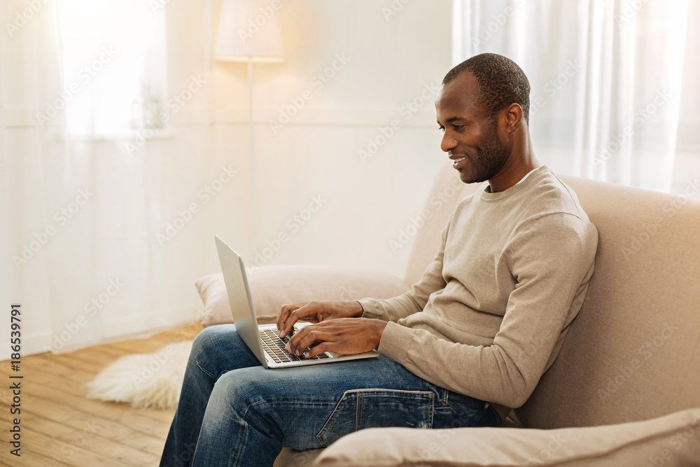 Freelancing. Happy smiling afro-american man typing on the computer and looking at the screen while sitting on the couch Freelancing. Happy smiling afro-american man typing on the computer and looking at the screen while sitting on the couch