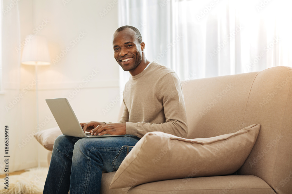 In high spirits. Content dark-eyed afro-american man grinning and working on the computer while sitting on the couch and a lamp and a carpet in the background In high spirits. Content dark-eyed afro-american man grinning and working on the computer while sitting on the couch and a lamp and a carpet in the background