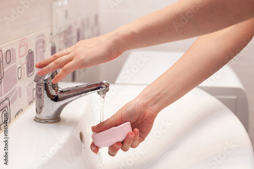 A woman takes a soap and opens a water tap to wash her hands in a white sink in the bathroom