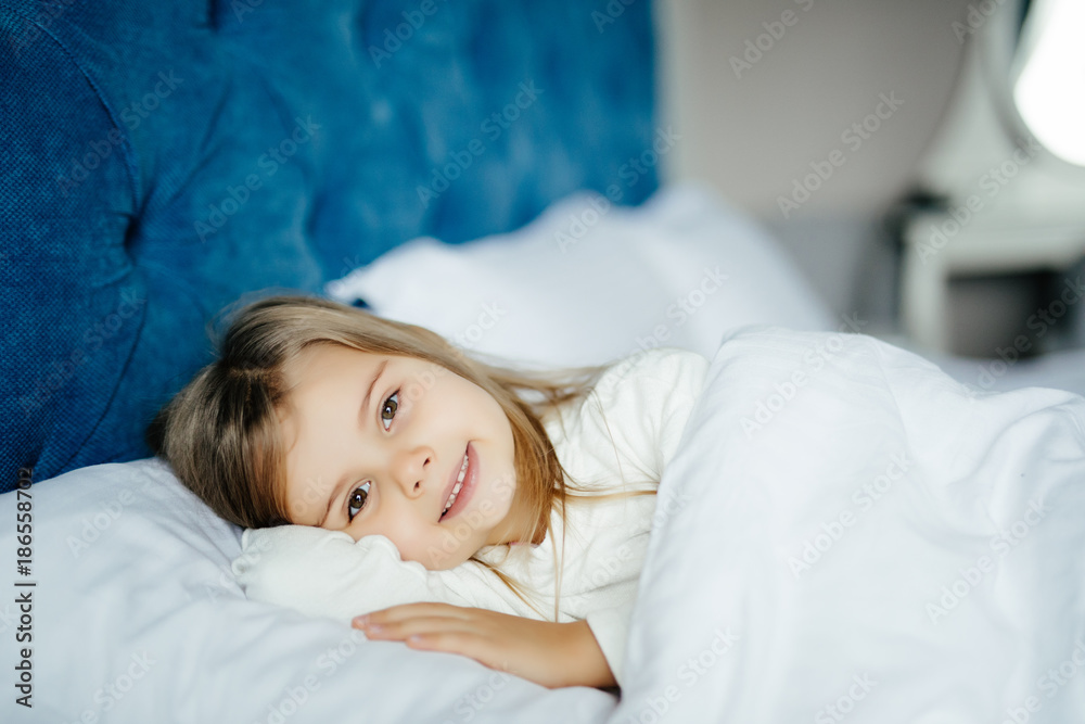 Close-up portrait of beautiful little kid lying on bed with hand under pillow, looking at camera at home Close-up portrait of beautiful little kid lying on bed with hand under pillow, looking at camera at home