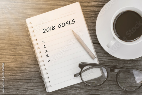 2018 Goals concept on notebook with glasses, pencil and coffee cup on wooden table.