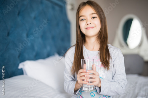 Beautiful little girl drinking water on white bed at home