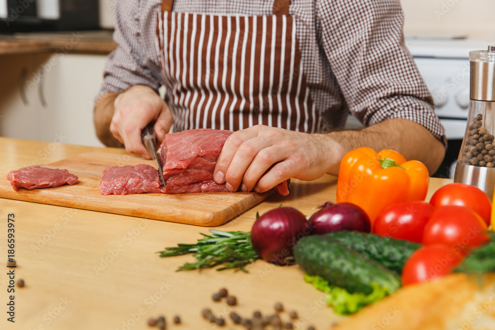 Close up caucasian young man in apron sitting at table with vegetables, cooking at home preparing meat stake from pork, beef or lamb, in light kitchen with wooden surface, full of fancy kitchenware.