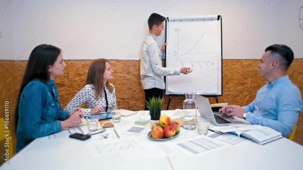 Young Enterpreneur is Presenting Company Growth Project to his Business Team in a Casual Office Room with Paperboard Young Enterpreneur is Presenting Company Growth Project to his Business Team in a Casual Office Room with Paperboard