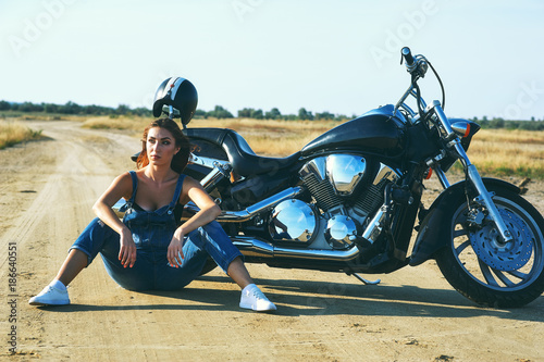 young woman in denim overalls , sitting on the ground near a motorcycle