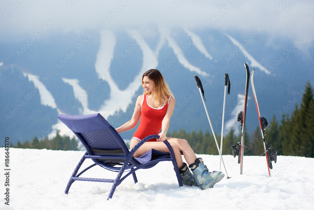 Attractive woman skier wearing red swimsuit, sitting on a blue deck chair near skis and poles at winter ski resort on the top of the hill. Mountains, ski slopes and forest on the background