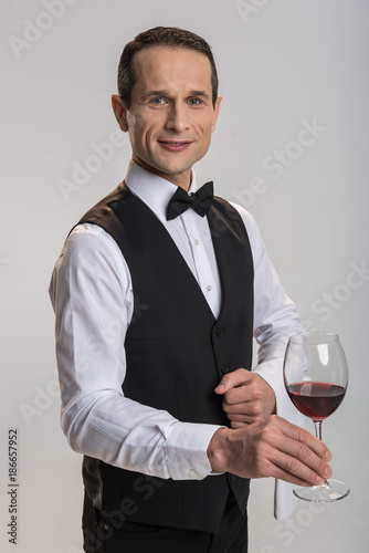Sophisticated wine. Pleasant gay male waiter smiling on background and gazing straight while holding glass of wine