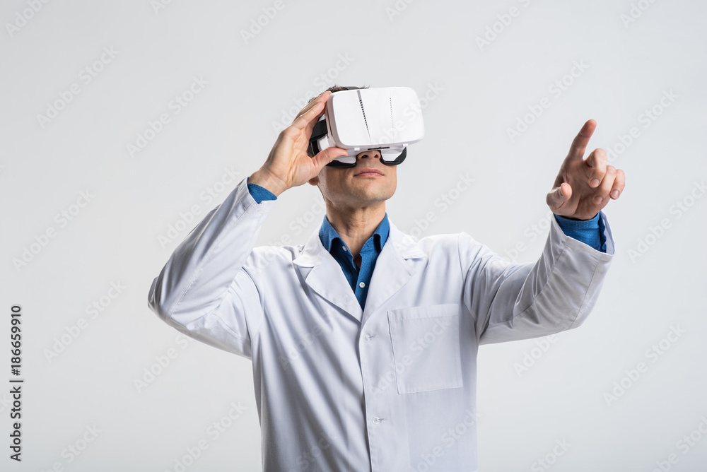 VR helps. Ambitious modern male doctor working in VR glasses while pointing with finger and reviewing system VR helps. Ambitious modern male doctor working in VR glasses while pointing with finger and reviewing system