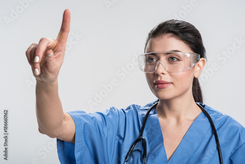 Technology inspires. Pensive satisfied female doctor  rising finger and smiling while gazing straight 