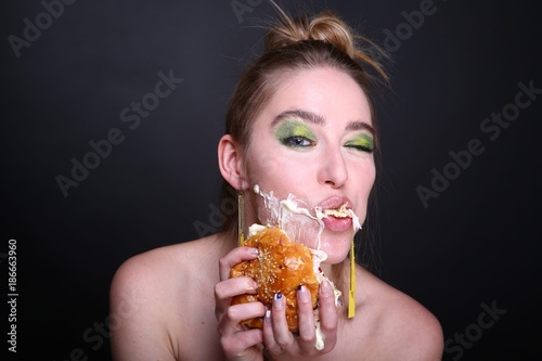 The woman is eating a giant hamburger with meat. Appetite lunch, delivery of food at home. Bright green fashionable make up eye shadows, yellow earrings. banner conceptual idea unhealthy food eating. photo