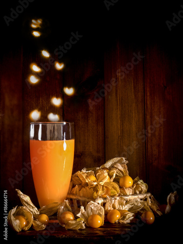 Cape gooseberries and a glass of cape gooseberry juice with heart shape light bokeh falling into glass on a wooden slab and wooden background in a dim room light, still life