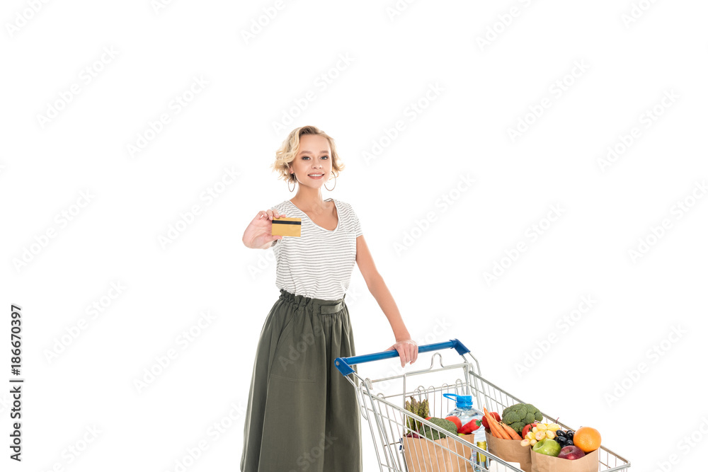 woman holding credit card and smiling at camera while standing with shopping trolley full of grocery isolated on white woman holding credit card and smiling at camera while standing with shopping trolley full of grocery isolated on white