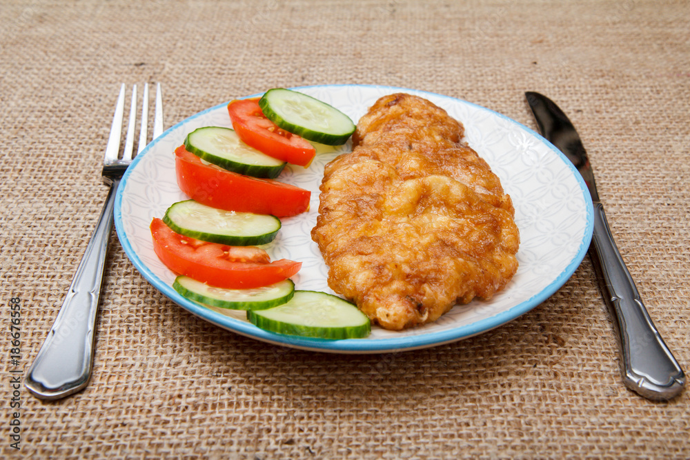 Plate with fried meat chop in egg batter and sliced tomato and cucumber with knife and fork Plate with fried meat chop in egg batter and sliced tomato and cucumber with knife and fork