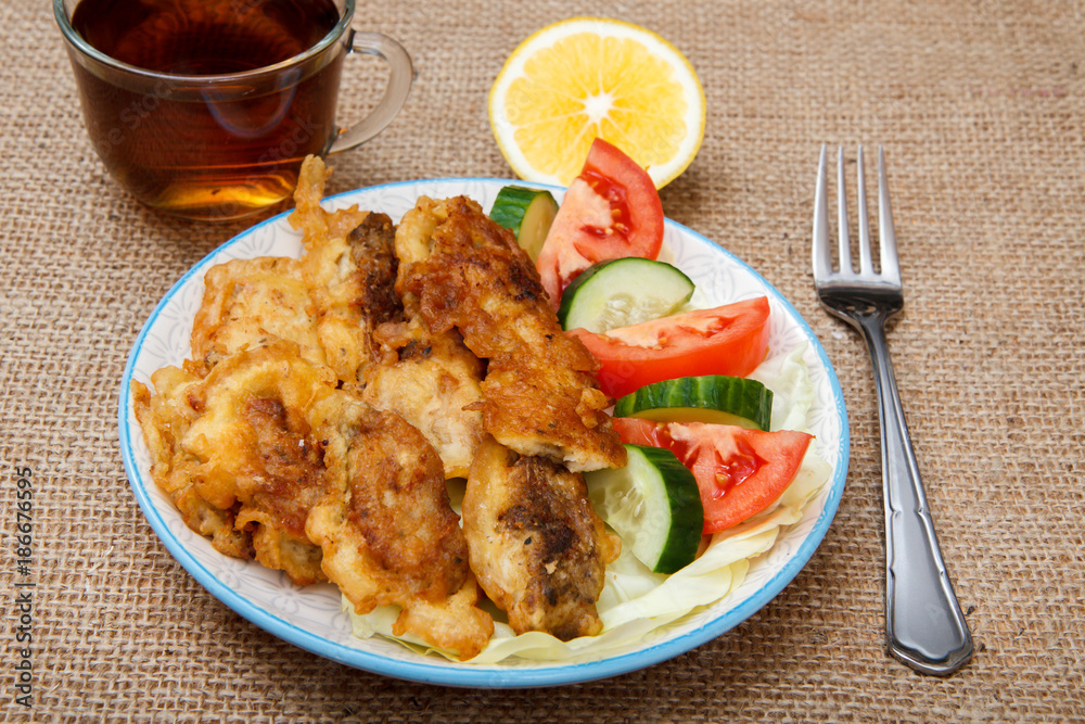 Plate with pieces of fried fish and freshly sliced tomatoes and cucumbers, cup of black tea with slice of lemon Plate with pieces of fried fish and freshly sliced tomatoes and cucumbers, cup of black tea with slice of lemon