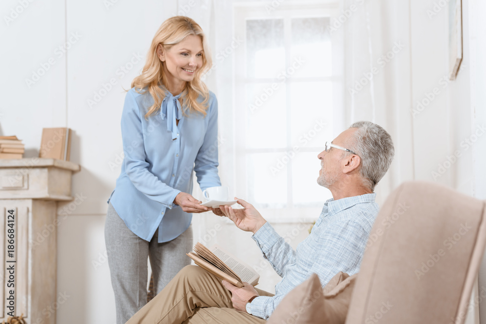 Obraz premium Mature man reading book while woman serves him tea in light room