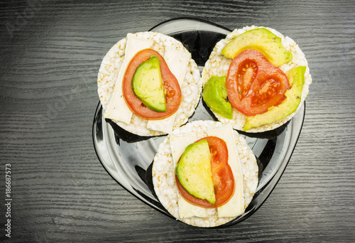 A black plate with prepared healthy and dietary sandwiches, a rice cake with cheese, tomato and avocado.