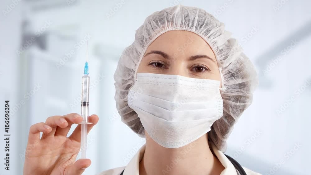 Professional female doctor in mask, cap and gloves in hospital room with disposable one-off syringe with transparent drug. Woman physician at work. Laboratory employee making scientific research Professional female doctor in mask, cap and gloves in hospital room with disposable one-off syringe with transparent drug. Woman physician at work. Laboratory employee making scientific research