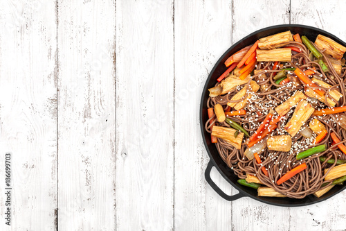 Delicious soba with tofu skin and vegetables in a cast iron pan on light wooden background, top view