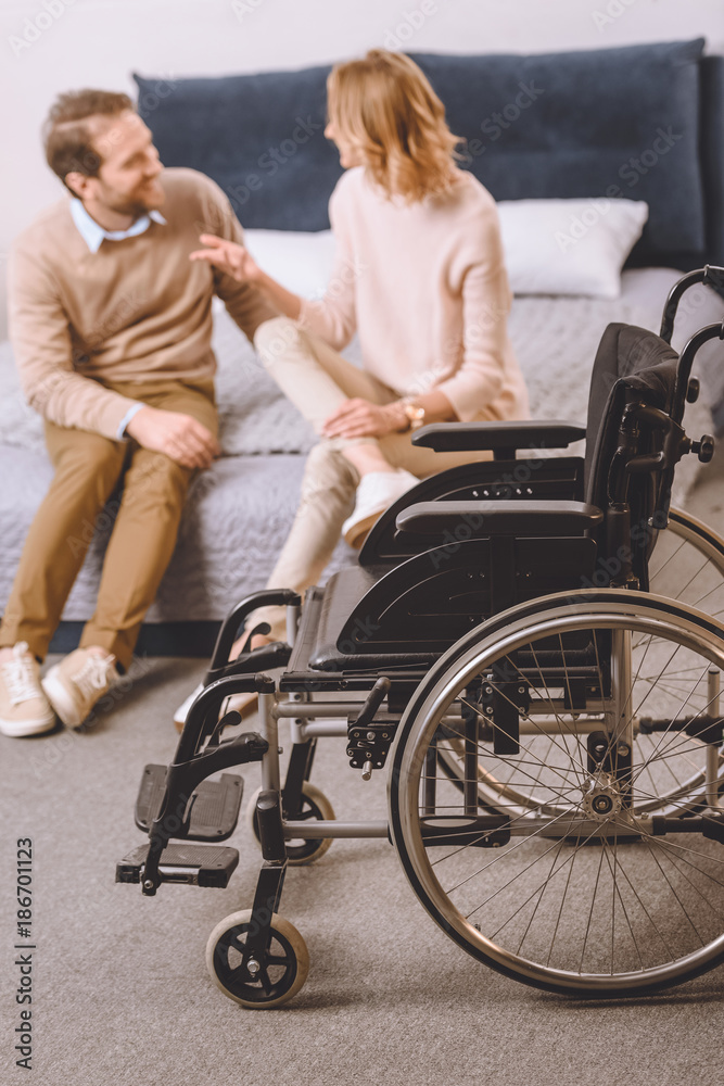 husband with disability and wife sitting and talking on bed with wheelchair on foreground husband with disability and wife sitting and talking on bed with wheelchair on foreground