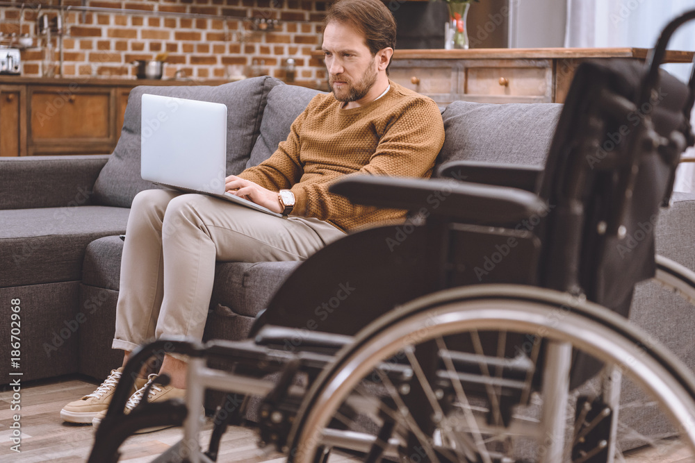 close-up view of wheelchair and disabled middle aged man using laptop on sofa at home close-up view of wheelchair and disabled middle aged man using laptop on sofa at home