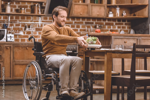 happy disabled man in wheelchair holding glass bowl of vegetable salad at home