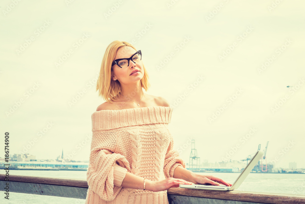 Eastern European Businesswoman traveling, working in New York, wearing off shoulder sweater, glasses, working on laptop computer by river in sunny spring day, looking up, thinking. Filtered effect..