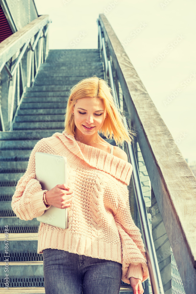 Eastern European College Student studying, working in New York, wearing knitted loose, off shoulder sweater, holding laptop computer, smiling, walking down stairs on campus. Filtered effect..