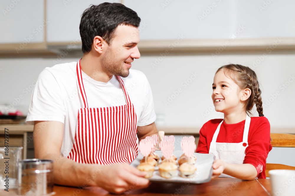 Indoor shot of handsome man wears striped apron and his small pretty female kid hold hand made cakes, rejoices successfully done work on kitchen, being in high spirit, happy spend time together.