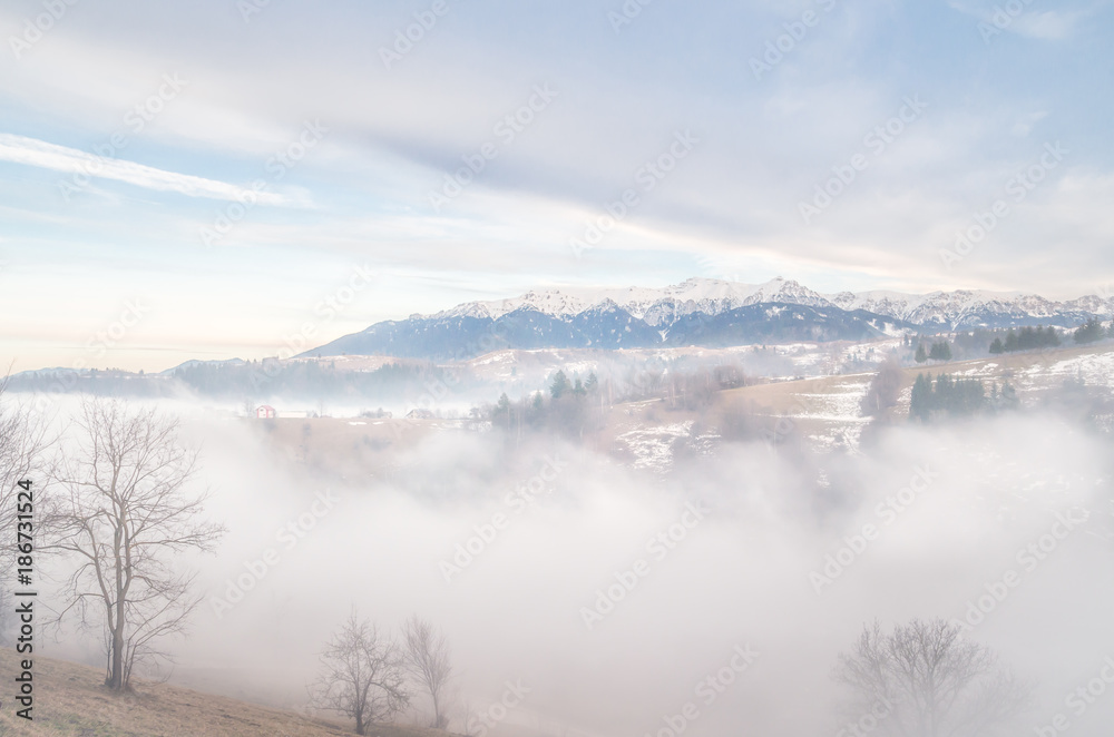 Winter landscape in the mountains. Rural landscape in the evening with fog and clouds on the sky. There are some houses in the image.