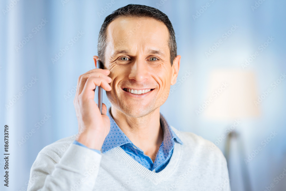Pleasant conversation. Charming middle-aged man in a blue shirt and a grey pullover talking on the phone and smiling in the camera Pleasant conversation. Charming middle-aged man in a blue shirt and a grey pullover talking on the phone and smiling in the camera