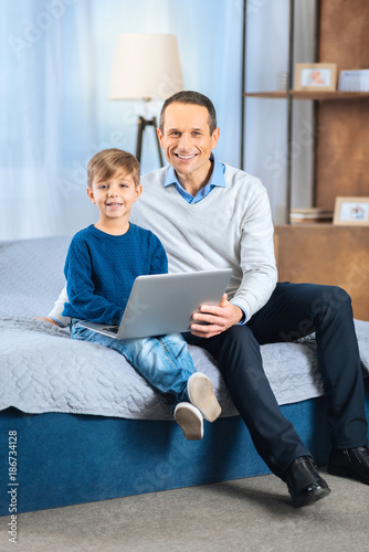 Happy users. Handsome young man sitting on the bed next to his little son and posing for the camera together with him while the boy holding a laptop in his lap