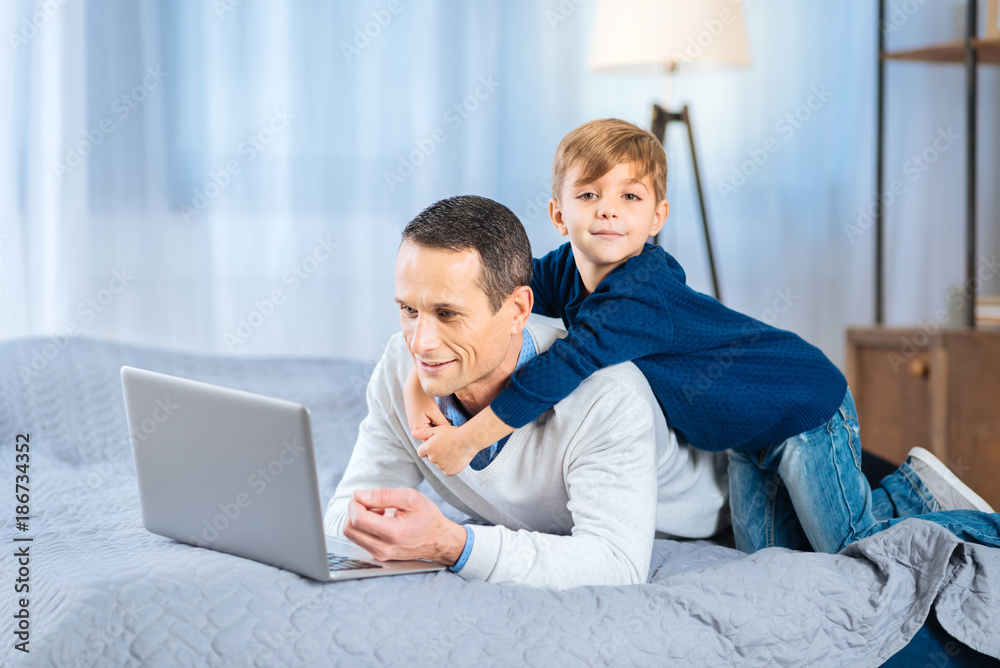 Gentle hug. Adorable little boy back-hugging his beloved father lying on the bed and working on the laptop Gentle hug. Adorable little boy back-hugging his beloved father lying on the bed and working on the laptop
