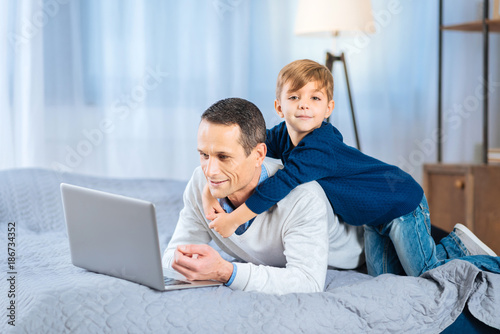 Gentle hug. Adorable little boy back-hugging his beloved father lying on the bed and working on the laptop