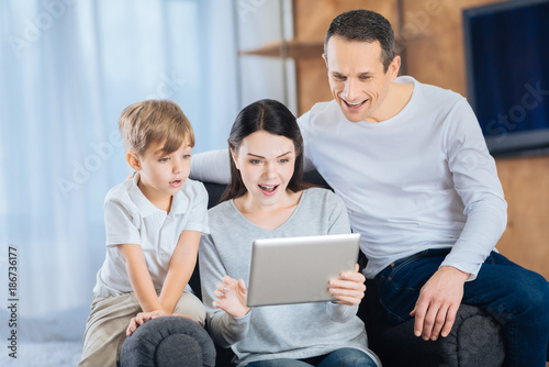 Surprising content. Pleasant young woman sitting in the armchair between her little son and husband and watching a video together with them