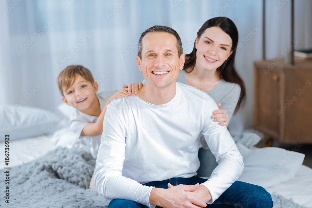 Head of the family. Charming young man sitting on the bed and smiling at the camera while his son and wife posing standing behind his back Head of the family. Charming young man sitting on the bed and smiling at the camera while his son and wife posing standing behind his back