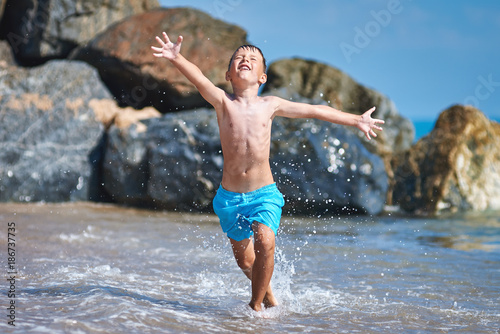 Cute Caucasian boy is running in the water along the sea shore. His arms are wide open.