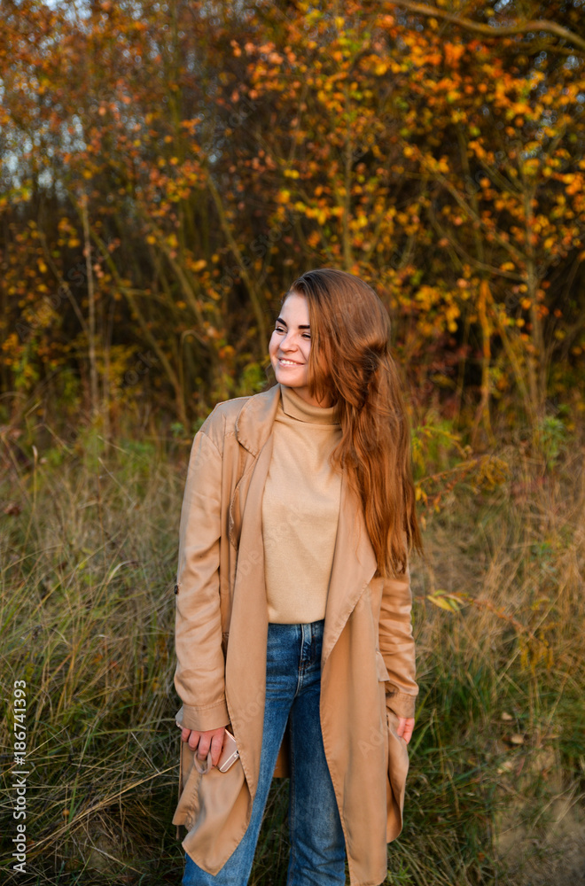 Stylish and beautiful red haired girl posing, walking in autumn forest among yellow grass. Woman with long hair, in jeans, blouse and brown coat, smiling, relaxing outdoors. Stylish and beautiful red haired girl posing, walking in autumn forest among yellow grass. Woman with long hair, in jeans, blouse and brown coat, smiling, relaxing outdoors.