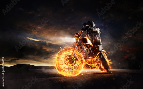 Fotografia Dark motorbiker staying on burning motorcycle in sunset light