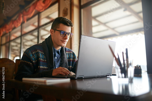 Handsome freelancer businessman working on laptop in cafe. Blogger man updating his profile in social networks with photos sharing with followers multimedia using notebook with wifi. Nerd in glasses.