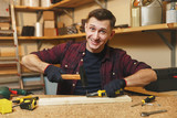 Handsome smiling caucasian young man in plaid shirt, black T-shirt, gloves hammering nails with hammer, working in carpentry workshop at wooden table place with piece of wood, different tools.