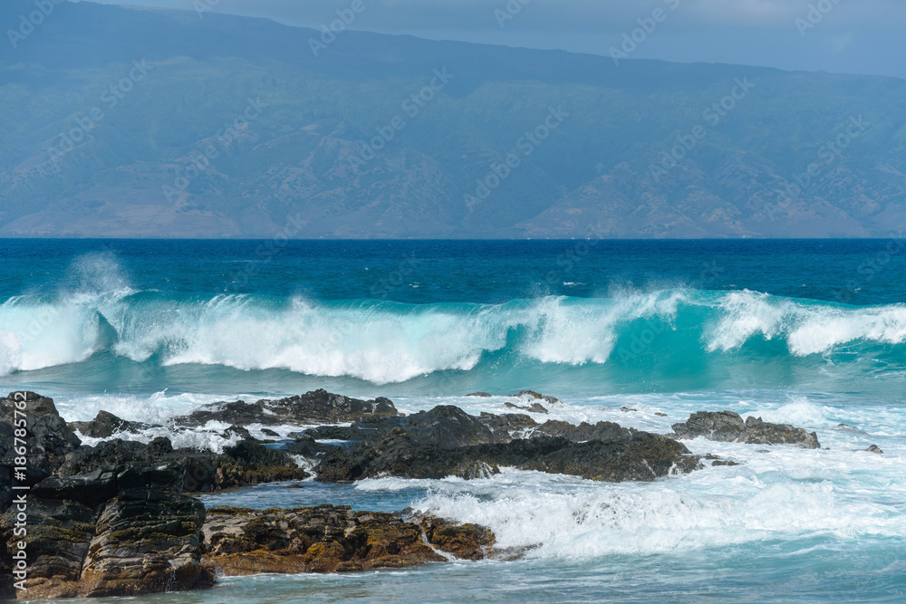 Breaking Waves - Big breaking blue waves rushing towards black rocky coast, with a tropical island in the background. Maui, Hawaii, USA.