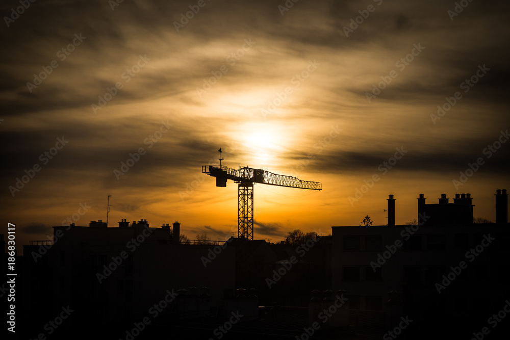 Naklejka premium Large industrial crane silhouetted at sunset against an orange sky rising above the rooftops of a city on the skyline
