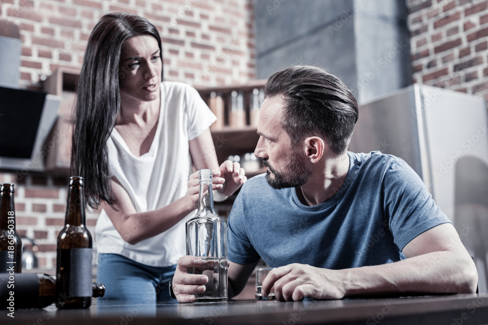 Unhappy marriage. Cheerless gloomy unhappy woman looking at her husband and asking him to stop drinking while trying to help him Unhappy marriage. Cheerless gloomy unhappy woman looking at her husband and asking him to stop drinking while trying to help him