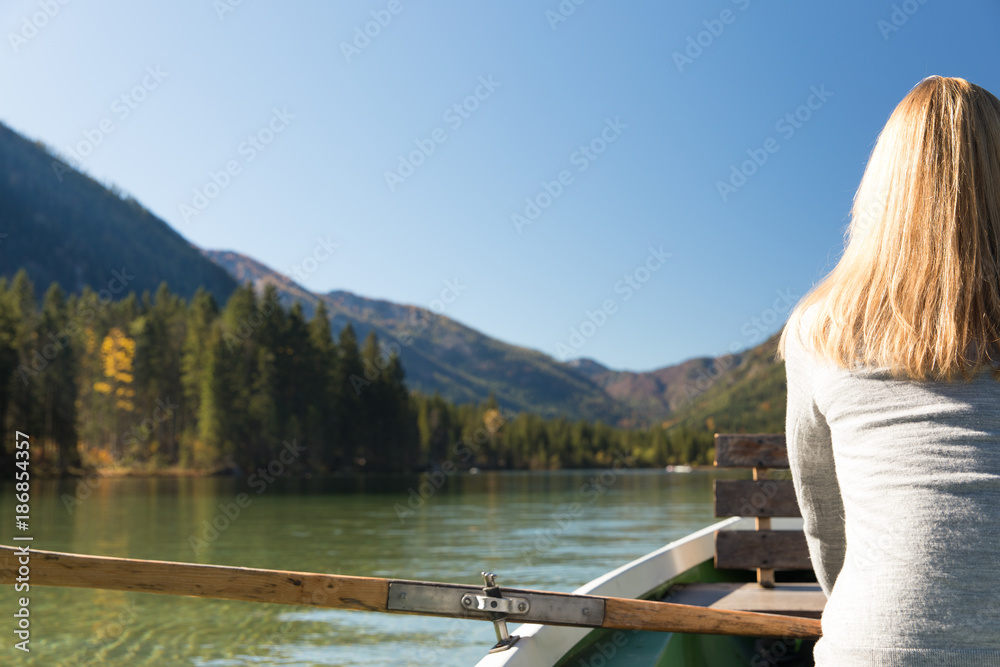 Junge, hübsche Frau rudert mit einem Ruderboot auf einem See in den Bergen Alpen an einem wunderschönen Herbsttag. Sie genießt die Sonne, liest ein buch und macht ein Selfie mit ihrem Handy Kamera