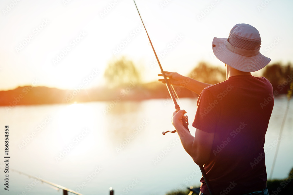 Young man fishing on a lake at sunset and enjoying hobby - obrazy, fototapety, plakaty 