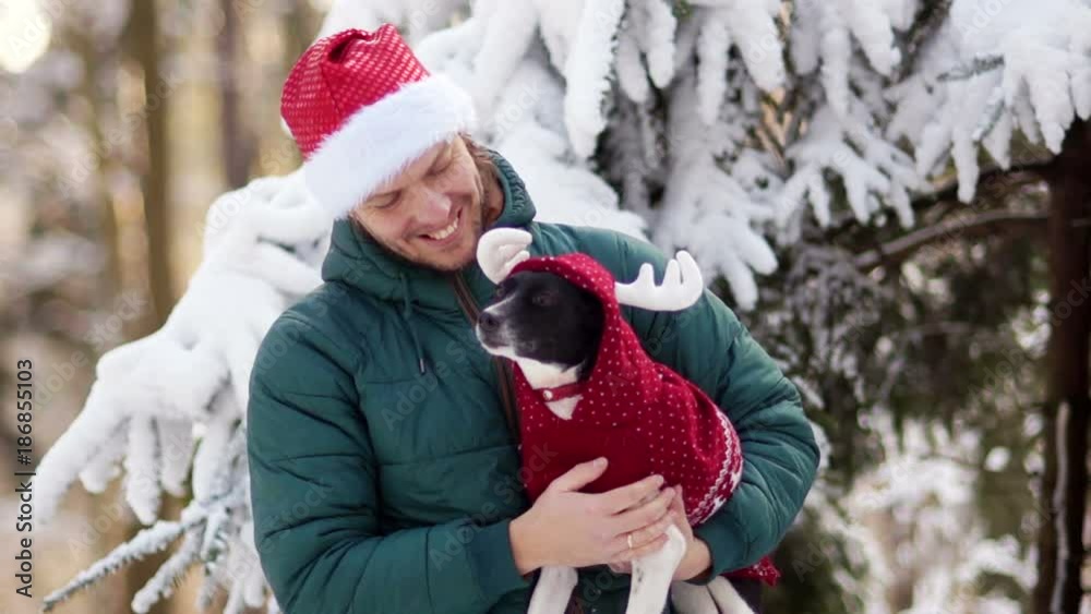 A dog in a New Year costume of a deer. Sits in the hands of the owner. A young man has a Santa hat on his head. In the snow-covered forest A dog in a New Year costume of a deer. Sits in the hands of the owner. A young man has a Santa hat on his head. In the snow-covered forest