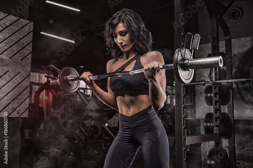 Young beautiful girl bodybuilder doing exercises in the gym using a barbell, exercises for the biceps, hard training, on a dark background in the smoke.