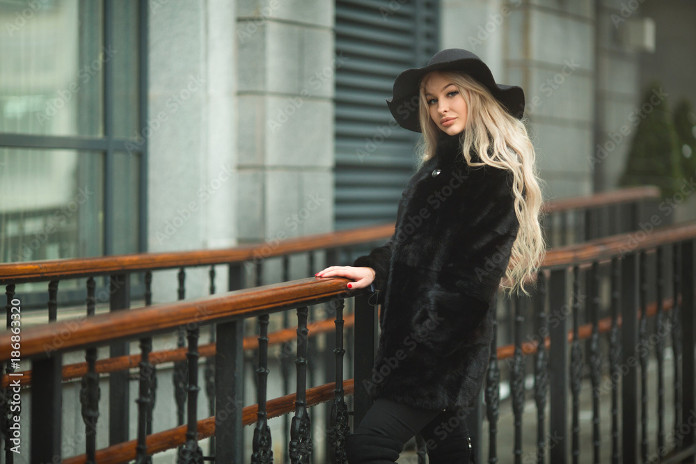 young beautiful girl in a black fur coat and hat on the street near the building