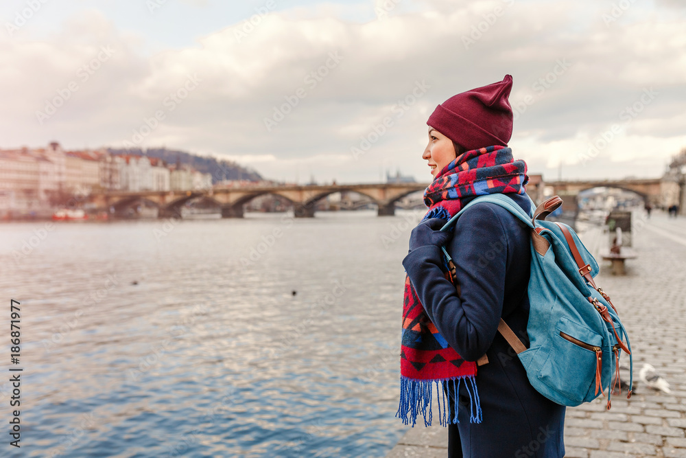 Young woman tourist in coat with backpack standing near bridge over the Vltava river in Prague