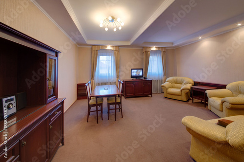 A light drawing room with upholstered furniture  a dining table  chairs  a sideboard and the TV set on a bedside table